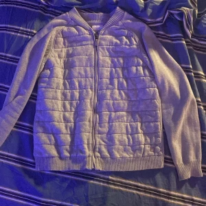 Grå kofta med dragkedja - Grå kofta med quiltad framsida och dragkedja hela vägen. Långärmad modell i mjukt stickat material, ribbade muddar och krage. Perfekt för lager-på-lager och chill dagar.