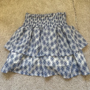 Vit och blå mönstrad kjol från H&M - Supersöt kort kjol från H&M i vitt och blått med ett snyggt grafiskt mönster. Kjolen har smock i midjan och dubbla volanger för extra volym. Tillverkad i lätt och luftig bomull, perfekt för varma dagar. Storlek 152 och ny skick💕