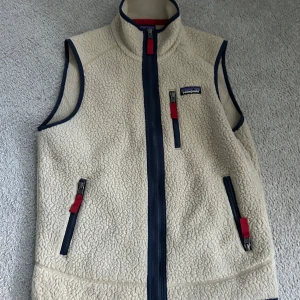 Beige fleeceväst från Patagonia XS - Säljer en beige fleeceväst från Patagonia i storlek XS. Västen har marinblå detaljer, röda dragkedjor och hög krage. Perfekt för lager-på-lager och outdoor-vibes. Två fickor framtill med dragkedja och klassisk Patagonia-logga på bröstet.