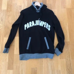 Svart hoodie från Parajumpers S - Svart hoodie från Parajumpers med gråa muddar och huva. Stor vit logga framtill och patch på ärmen. Känguruficka och snörning i huvan. Perfekt för dig som gillar streetstyle och vill ha något bekvämt och snyggt.