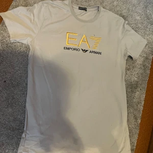 Beige EA7 Emporio Armani t-shirt - Snygg beige t-shirt från EA7 Emporio Armani med gul broderad logga på bröstet. Klassisk rund hals och korta ärmar. Perfekt för dig som gillar stilrena märkesplagg med en exklusiv touch.