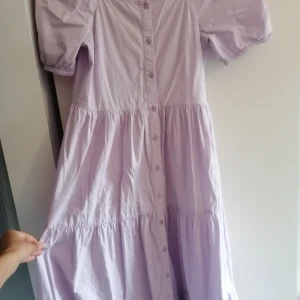 Ljuslila långklänning från Lindex - Supersöt ljuslila långklänning från Lindex med puffärmar och knappar hela vägen framtill. Klänningen har en luftig passform och är sydd i mjuk bomull, perfekt för varma dagar. Volangdetaljer ger extra flow och en romantisk vibe. 💞🌺                                                   Klänningen har bara använts bara ett fåtal gånger. Det har en liten blå fläck under en knapp (syns knappt). 