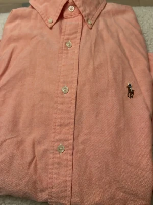 Rosa skjorta från Ralph Lauren - Klassisk rosa skjorta från Ralph Lauren med button-down krage och broderad logga på bröstet. Skjortan har vita knappar framtill och är tillverkad i mjuk bomull. Perfekt för dig som gillar stilrena och tidlösa plagg.