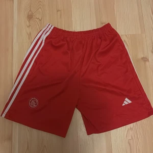 Röda Ajax fotbollsshorts Adidas - Säljer ett par röda fotbollsshorts från Adidas med Ajax-logga på ena benet och klassiska vita ränder längs sidorna. Shortsen har elastisk midja och är gjorda i ett lätt syntetmaterial som passar perfekt för träning eller match.