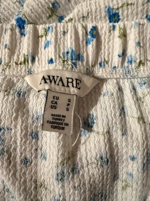 Blommig långkjol från AWARE - Säljer en vit långkjol från AWARE med små blå blommor och gröna detaljer. Kjolen har en rak passform, elastisk midja och en slits på sidan för extra rörelsefrihet. Materialet är strukturerat och känns mjukt mot huden. 