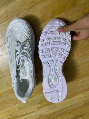 Nike Air Max 97 vita sneakers - Nike Air Max 97 sneakers i helvit färg med snygga vågiga linjer och meshdetaljer. Skorna har snörning, platt sula och rund tå. Ikonisk Air Max-bubbla i sulan för extra komfort. Perfekt för dig som gillar stilrena och sportiga sneakers.