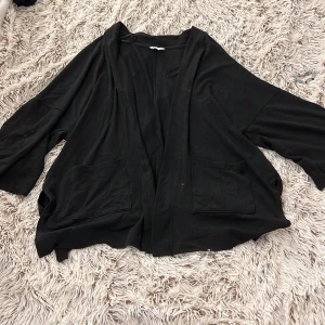 Svart kofta med fickor från Cubus - Svart, mjuk och luftig kofta från Cubus i storlek M. Koftan har trekvartsärmar, två stora fickor framtill och en avslappnad passform. Perfekt att slänga på sig över en t-shirt eller linne när det är lite kyligare ute.