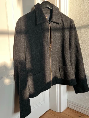 Gråsvart kort jacka i ull - Jacka i ull med tunt svart foder, rak modell. Svart- mörkgrå färg. Jackan är sent 90-tal från Lemon med diskret  fiskbensmönstret tyg, tillverkad i 100% virgin ull. Klassisk krage, två framfickor och en cool guldfärgad dragkedja med ring. Ett stilrent och tidlöst plagg -  ny vintage. Passar strl S/M. 