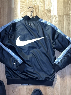 Nike bomberjacka går använda båda sidor  - Nike bomberjacka med stor vit Swoosh-logga på ryggen och mindre loggor på bröstet och ärmarna. Jackan är svart med silverfärgade paneler längs ärmarna och har ribbade muddar. Framsidan har dragkedja och fickor. Materialet är polyester och jackan har quiltad insida. Jackan är i bra skick inga skador eller skrynklig osv väldigt fin jacka som passar de mesta st s/m