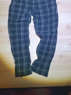 Mörkgröna rutiga mjukisbyxor - Säljer ett par mörkblå mjukisbyxor med vitt och grått rutmönster. Byxorna har en avslappnad passform och är perfekta för chill dagar hemma. Materialet är mjukt och bekvämt, vilket gör dem sköna att bära länge. Passar dig som gillar en casual stil.