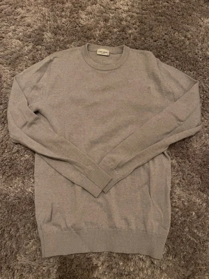 Grå stickad tröja från John Henric - Snygg grå stickad tröja från John Henric i storlek S. Tröjan har rund halsringning och långa ärmar med ribbade muddar vid ärmslut och nederkant. Perfekt för lager-på-lager och enkel att matcha med jeans eller chinos.