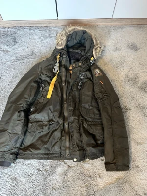 Parajumper mörkgrön vinterjacka  - Vinterjacka från Parajumper i mörkgrönt med gul detaljrem, päls på huvan och flera fickor. Jackan har dragkedja, knappar och patch på ärmen. Materialet är polyester och jackan är fodrad för extra värme. Perfekt för kalla dagar.