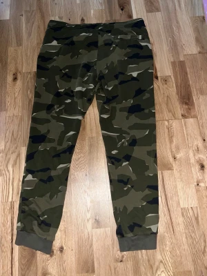 Nike camo mjukisbyxor med snörning - Säljer ett par mjukisbyxor från Nike med grön camouflage-mönster och grå muddar vid bensluten. Byxorna har snörning i midjan och en vit Nike-logga på ena sidan. Perfekta för chill eller streetwear.