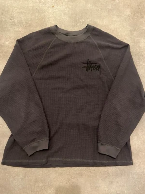 Svart långärmad tröja från Stüssy - Svart långärmad tröja från Stüssy med stort logotryck på ryggen och mindre logga på bröstet. Tröjan har en grov, rutmönstrad struktur och ribbade muddar vid ärmslut och hals. Perfekt för dig som gillar streetwear och vill ha något som sticker ut.