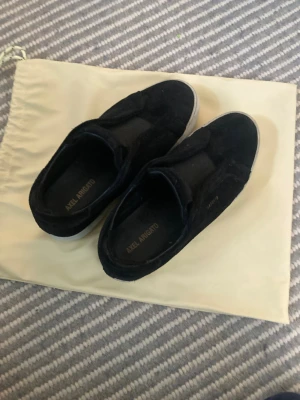 Svarta sneakers från Axel Arigato - Säljer ett par svarta sneakers från Axel Arigato med klassisk rund tå och platt vit sula. Skorna är tillverkade i mocka och har en stilren design utan synliga snörningar. Perfekt för dig som gillar minimalistisk och modern stil.