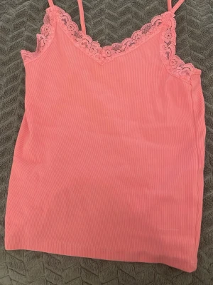 Rosa ribbad linnetopp med spets - Säljer en ljusrosa linnetopp med smala axelband och vacker spetsdetalj längs v-ringad hals. Toppen är ribbad och har en mjuk, feminin look som passar perfekt till sommaren. Materialet känns lätt och bekvämt mot huden.