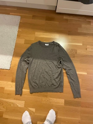 Grå stickad tröja från Lindbergh - Säljer en enkel och stilren grå stickad tröja från Lindbergh i storlek L. Tröjan har rund hals, ribbade muddar och lång ärm. Perfekt att slänga på sig när det är lite kyligt ute. Passar dig som gillar klassisk och clean stil.