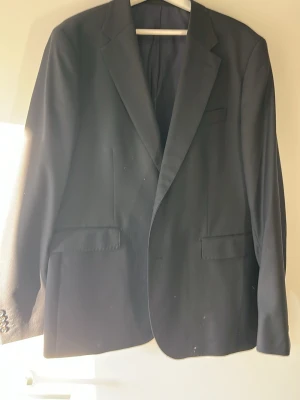 Svart kavaj från These Glory Days - Stilren svart kavaj från These Glory Days i storlek 58. Klassisk modell med snygga slag, tre framfickor och fyra knappar vid ärmslutet. Materialet känns mjukt och har en lätt glans. Perfekt för dig som vill ha en clean och tidlös look.