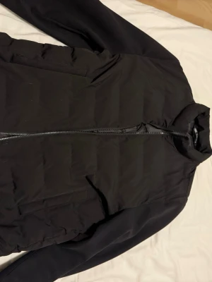 Svart pufferjacka från Zara Man - Svart pufferjacka från Zara Man med stilren design och raka linjer. Jackan har quiltad framsida, två fickor och hel dragkedja. Utsidan är i polyester och insidan har ett glansigt foder. Perfekt för kyliga dagar och enkel att matcha med din outfit.