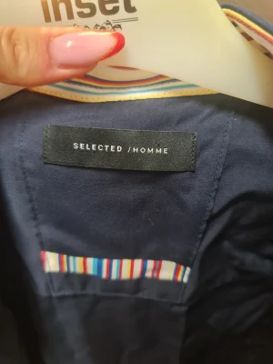 Mörkblå skjorta Selected Homme - Snygg mörkblå skjorta från Selected Homme med färgglada ränder på insidan av knappslån och kragen. Skjortan har röda knapphål och svarta knappar, vilket ger en cool kontrast. Perfekt för dig som vill ha något extra i garderoben.