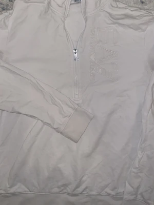 Vit EA7 långärmad tröja med half zip - Säljer en stilren vit långärmad tröja från EA7 Emporio Armani med half zip framtill. Tröjan har ett stort, upphöjt EA7-logotryck på bröstet och är gjord i mjuk bomull. Perfekt för dig som gillar clean och sportig look.