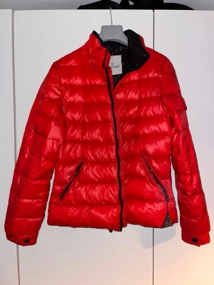 Röd Moncler pufferjacka, storlek 2 - Säljer en riktigt snygg röd pufferjacka från Moncler. Jackan har svart innerfoder, dragkedja framtill och två fickor med dragkedja. Klassisk Moncler-logga på ärmen och cool quiltad design. Perfekt för kalla dagar när du vill sticka ut lite extra.