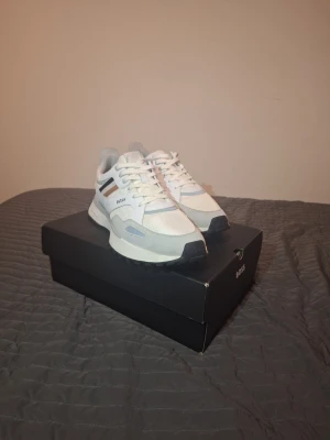 Vita sneakers från BOSS - Säljer ett par fräscha vita sneakers från BOSS med snygga detaljer i grått, svart och beige. Skorna har snörning, rund tå och platt sula. Materialmix av syntet och skinn med meshpaneler på ovansidan för extra komfort. Perfekt för dig som gillar stilrena och moderna sneakers.