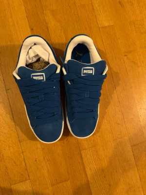 Blå Puma Suede XL sneakers - Säljer ett par fräscha Puma Suede XL sneakers i blå mocka med vita detaljer och guldlogga på sidan. Skorna har rund tå, platt sula och klassisk snörning. Insidan är vit och yttersulan är vit med struktur. Perfekta för dig som gillar stilrena och sportiga sneakers.