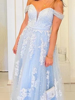 Ljusblå långklänning med spets - Magisk ljusblå långklänning med offshoulder och v-ringad urringning. Klänningen har vackra vita spetsdetaljer och blommigt mönster över hela tyget, samt små pärlor som ger extra lyxig känsla. Tillverkad i ett tunt, svepande material som ger en drömmig siluett.