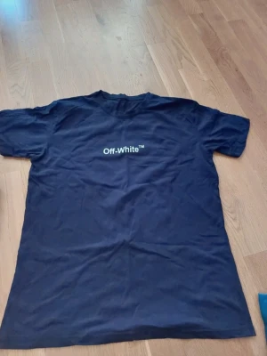 Off-White x Kaws mörkblå t-shirt - Säljer en mörkblå t-shirt från Off-White med Kaws-motiv på ryggen och Off-White-logga på bröstet. T-shirten är kortärmad och har en normal passform. Materialet är mjuk bomull och trycket är detaljerat med grafiskt motiv.