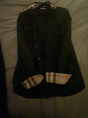 Svart skjorta Burberry - Svart skjorta från Burberry med klassiska rutiga detaljer på insidan av kragen och ärmsluten. Snygg och stilren design som sticker ut. Storlek L. (xl men sitter som L/M)