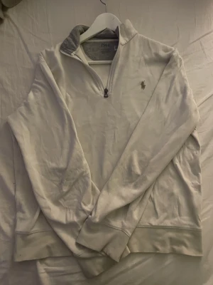 Vit half zip tröja från Polo Ralph Lauren - Vit långärmad tröja från Polo Ralph Lauren med grå insida på kragen och klassisk broderad logga på bröstet. Tröjan har half zip-dragkedja och är tillverkad i mjuk bomull, perfekt för chill dagar och snygg layering.för de som är 170 ungefär 