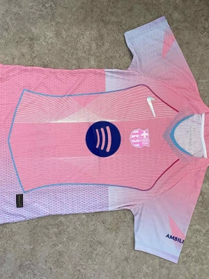 Ingen sko på bilden - Bilderna visar en rosa och lila FC Barcelona fotbollströja med Nike-logga, Spotify och UNHCR tryck, men inga skor finns med.