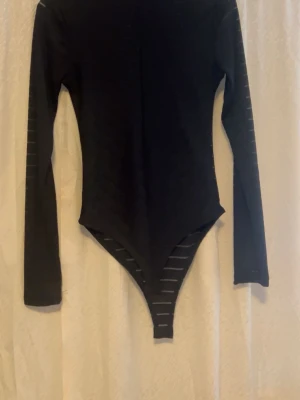 Svart randig långärmad bodysuit - Säljer en svart bodysuit med lång ärm och coola genomskinliga ränder. Toppen har en tight passform och är tillverkad i ett stretchigt syntetmaterial som sitter snyggt mot kroppen. Perfekt för att styla med jeans eller kjol för en trendig look.