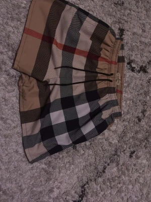 Burberry - Säljer ett par snygga rutig shorts från Burberry med klassiskt beige, svart och rött mönster. De har elastisk midja med snörning och är gjorda i mjuk polyester. Perfekta för sommarens alla häng och chill dagar.