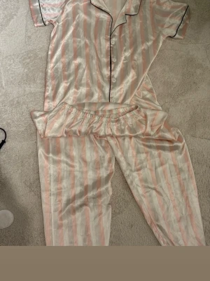 Randig satinväst i ljusrosa och vit - Säljer en glansig väst i satin med vertikala ränder i ljusrosa och vitt. Västen har klassisk krage, knappar framtill och svarta kantdetaljer. Perfekt för dig som gillar en mjuk och lyxig känsla. Passar dig som vill sticka ut med en söt och fräsch look.