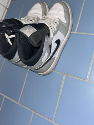 Nike Air Jordan 1 High Grå/Vit - Nike Air Jordan 1 High i grått, vitt och svart med klassisk Jordan-logga på sidan och tungan. Skorna har snörning, perforerad tå och rund tå. Materialet är skinn och syntet, med högt skaft och platt sula. Snygg och ikonisk modell, den är använd par gånger. Så riktigt bra skick 