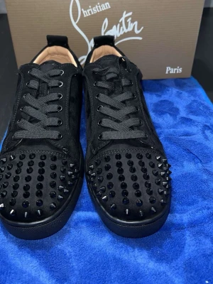 Christian Louboutin svarta sneakers med nitar - Svarta sneakers från Christian Louboutin i mocka med svarta nitar på tån och klassisk röd sula. Skorna har snörning och en rund tå, vilket ger en edgy och lyxig vibe. Perfekta för dig som vill sticka ut med exklusiva detaljer och streetstyle.