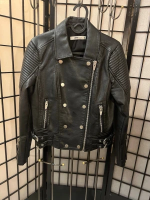 Svart skinnjacka från Gina Tricot, typ oanvänd - Snygg svart skinnjacka med silvriga dragkedjor och knappar. Jackan har quiltade detaljer på axlarna, två fickor med dragkedja och spännen i midjan. Klassisk bikerstil med coola detaljer och rak passform. Perfekt för dig som vill ha en edgy look.