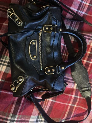 Svart handväska i skinn - Säljer en svart handväska med snygga guldfärgade metalldetaljer och dragkedja framtill. Väskan är tillverkad i äkta skinn och har både handtag och axelrem. Defekter syns i sista bilden 