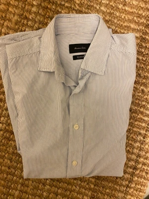 Randig skjorta från Massimo Dutti - Snygg skjorta från Massimo Dutti i vitt och ljusblått med tunna ränder. Tillverkad i 100% bomull, har klassisk krage, långa ärmar och knappar framtill. Perfekt för dig som gillar stilrena och fräscha plagg.