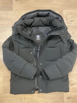 Svart puffer jacks från Canada Goose XS - Säljer en svart puffer jacka  från Canada Goose i storlek XS passar även S. Jackan har en stor huva, dragkedja med logotyp, tryckknappar och två sidofickor. Fylld med dun och fjädrar för extra värme, perfekt för kalla dagar. Klassisk pufferstil med stilren look.