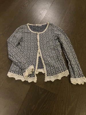 Blåvit stickad kofta med mönster - Supersöt blåvit stickad kofta från RosenRosen med detaljerat geometriskt mönster. Koftan har rund hals, lång ärm och knappar framtill. Fina virkade kanter och volangdetaljer vid nederkant och ärmslut ger extra charm. Perfekt att slänga på över en topp! Köpt sacond hand snören bak borta men dem behövs ej😀