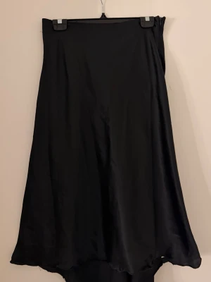 Svart långkjol i satin  - Säljer en svart långkjol i mjukt satinmaterial med diskret slits och vågig nederkant som jag sytt om själv. Den är kortare framtill och längre där bak. 