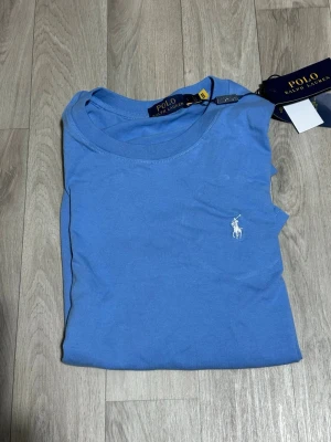 Blå t-shirt  - Snygg blå t-shirt från  med klassisk rund hals och korta ärmar. T-shirten har det ikoniska broderade polospelaren-loggan i vitt  ej äkta på bröstet. Tillverkad i mjuk bomull som känns skön mot huden. Perfekt för en chill och stilren look.