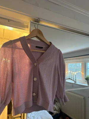 Glittrig lila kofta från Stockh LM - Säljer en glittrig lila kofta från Stockh LM med tre blommiga knappar framtill. Koftan har en croppad passform och puffiga trekvartsärmar.