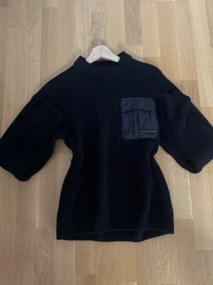 Svart stickad tröja Jack & Jones - Svart stickad tröja från Jack & Jones Core med en stor ficka i nylonmaterial på bröstet. Tröjan har ribbad struktur, rund hals och långa ärmar. Perfekt för dig som gillar streetwear och vill ha något som sticker ut med cool detalj framtill.