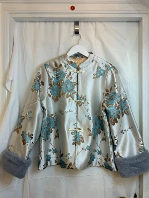 Elegan silk jacket - Elegant silk jacket with beautiful floral jacquard pattern and traditional Chinese knot buttons. Soft, slightly shiny surface with a luxurious feel. The sleeves feature delicate faux-fur cuffs, adding a unique and stylish touch.  Easy to style for both everyday wear and special occasions – looks great with jeans or skirts.  Size: 3XL (Chinese sizing, smallest size in this model is XL) Measurements: • Bust: 116 cm • Shoulder width: 41 cm • Length: 62 cm