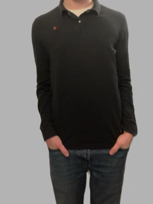 Svart långärmad skjorta Morris S - Svart långärmad skjorta från Morris i storlek S. Klassisk pikémodell med krage och knappar framtill. Diskret Morris-logga broderad på bröstet. Skjortan är gjord i mjuk bomull och har en normal passform som funkar till jeans eller chinos.