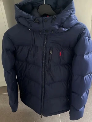Mörkblå pufferjacka från Polo Ralph Lauren - Snygg mörkblå pufferjacka från Polo Ralph Lauren med huva och dragkedja framtill. Jackan har en broderad röd logga på bröstet och flera praktiska fickor med dragkedja. Den har en lagning på ryggen cirka 6cm lång men annars inga defekter! 